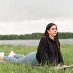 Woman lying on a yoga mat in a grassy field by a lake, promoting relaxation and wellness.